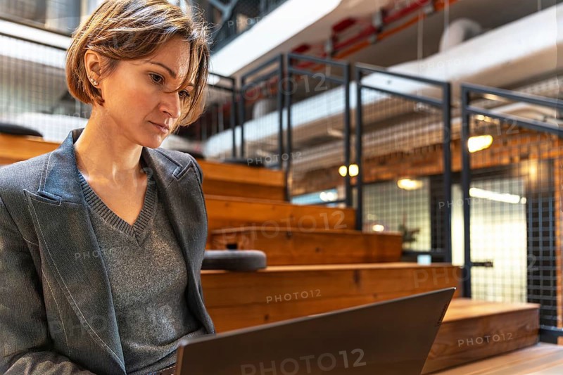 Professional woman in blazer with laptop on wooden steps in coworking space