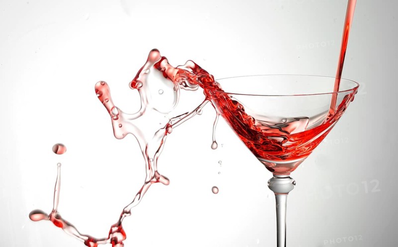 A powerful flood of red liquid pours into a cocktail glass and spills over, studio shot against a white background