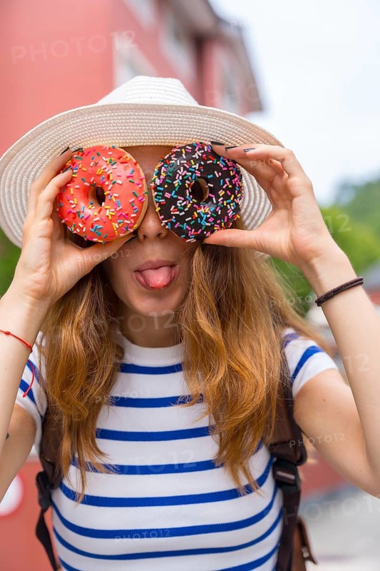 Young woman wearing a hat and striped shirt, holding two sprinkle covered donuts over her eyes and sticking out her tongue, enjoying a fun moment outdoors with sweet treats