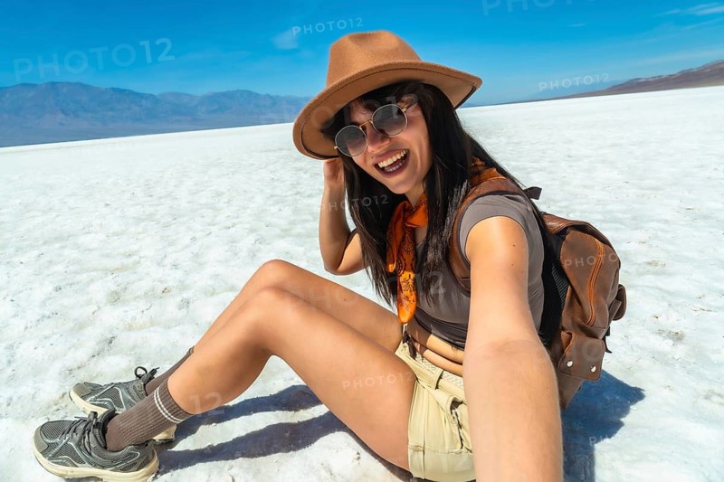 Woman wearing a hat and sunglasses, smiling and capturing a selfie while sitting on the vast white salt flats of badwater basin in death valley national park under a bright blue sky