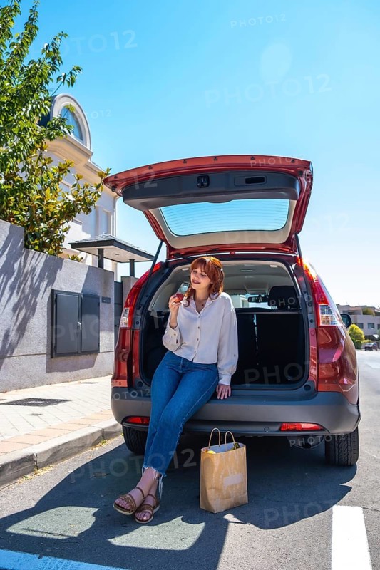 Woman sitting on open suv trunk eating an apple, relaxed after grocery shopping on a sunny day, casual jeans and white shirt, brown paper bag of fresh produce nearby