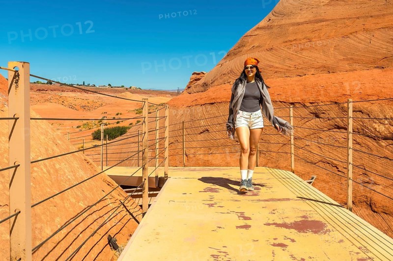 Woman in casual attire walking on a viewing platform through the impressive red sandstone rock formations and desert landscape of biidi the arch cave under a clear blue sky