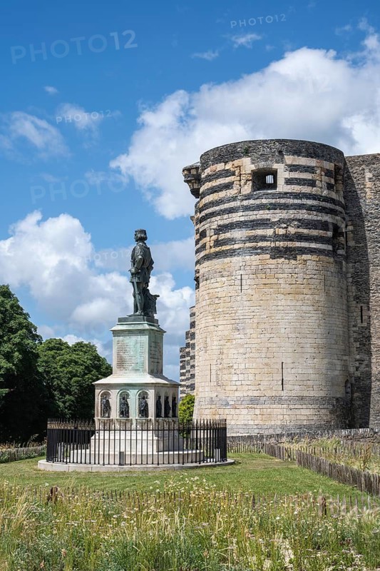 Bronze statue of King René on a decorated stone base in the foreground in front of one of the mighty, striped defensive towers of the historic Château d'Angers castle complex made of slate and limestone under a blue summer sky with loose clouds, Angers, Maine-et-Loire, Pays de la Loire, France