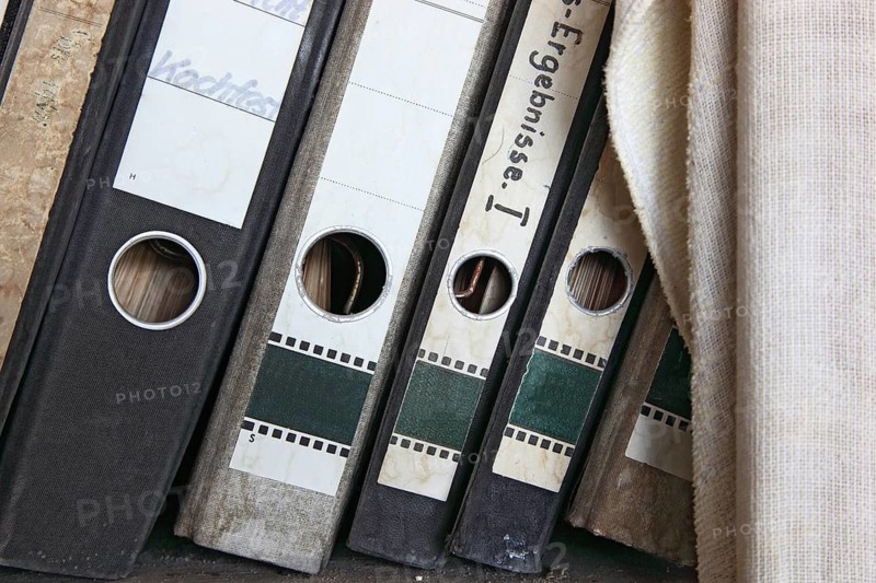 Office scene around 1982, lost place, abandoned location, classic file folders on a wooden shelf, some of the folders are labelled in detail by hand and provide information about their contents, mixed up, dirty