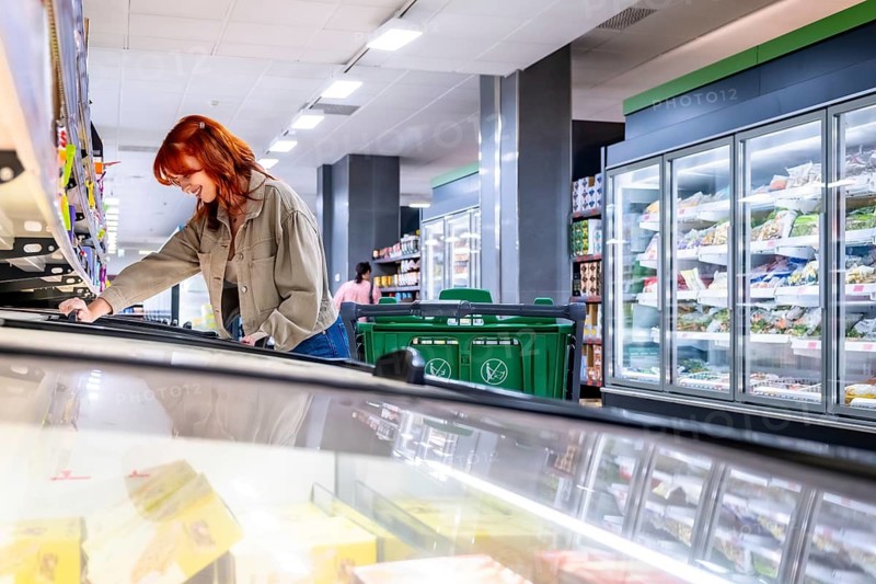Redhead woman smiling while pushing a green shopping cart and reaching into a supermarket freezer to choose frozen food in a modern, bright grocery aisle