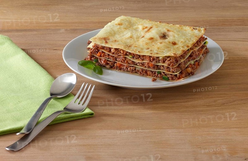 Lasagna on white plate decorated with basil leaf, fork, spoon and napkin next to it, on wooden table, studio shot