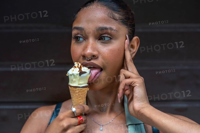 Young woman with a septum piercing enjoying a sweet dessert, licking a melting ice cream cone with pleasure on a warm summer day, standing against a dark background