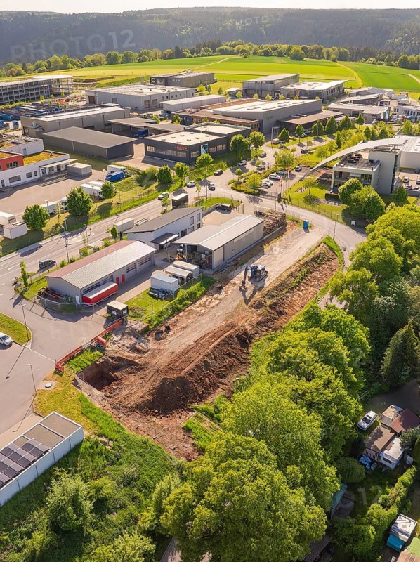 An aerial view of a construction site next to industrial buildings, surrounded by green trees and fields under bright sunshine, Parkhaus Bau, Calw, Germany