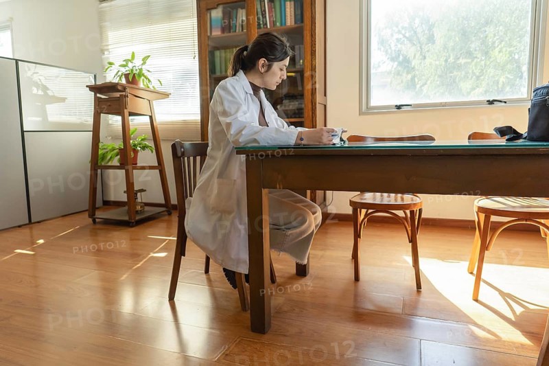 Young female scientist in white lab coat sitting at a large wooden table in a warm sunlit room with indoor plants, wooden bookcase and plant stand in the background