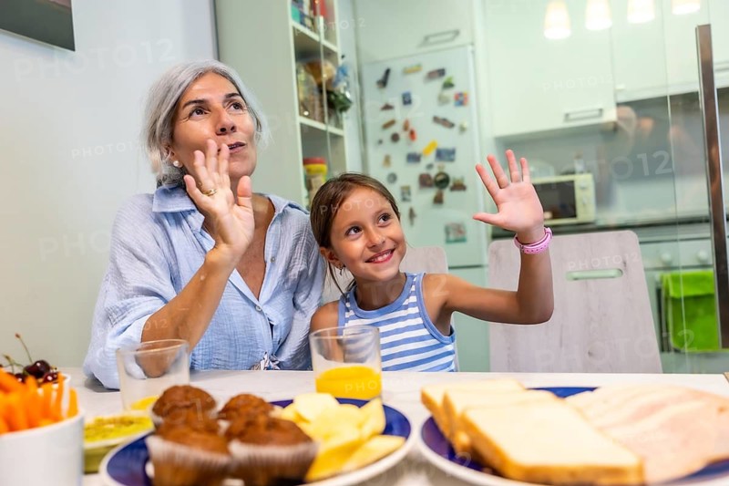 Older woman and young girl sitting at a kitchen table, smiling and waving during a family meal together, sharing a happy moment at home