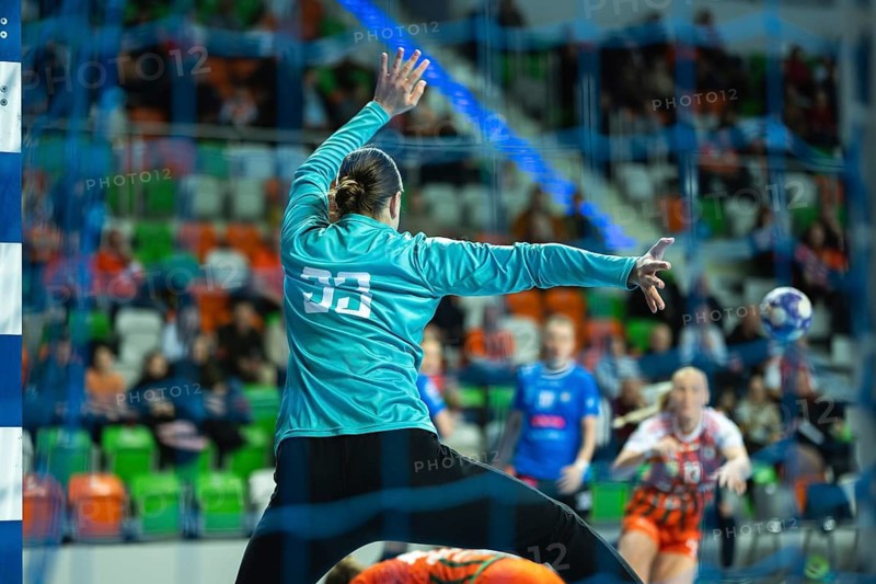 Professional female handball goalkeeper in a turquoise jersey performing a dynamic jump save during an intense indoor match in a crowded arena in Lubin, Poland