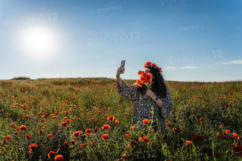 Woman taking selfie in blooming poppy (Papaver rhoeas) field while wearing floral crown and holding bouquet under bright summer sun