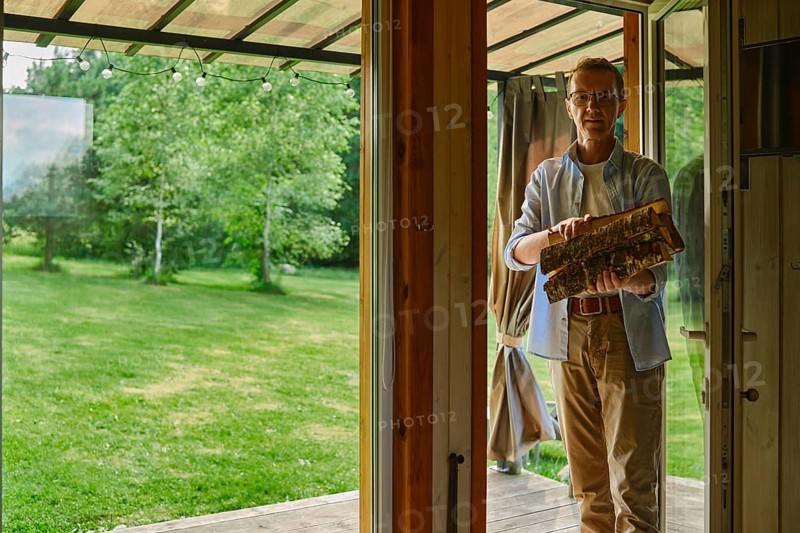 Middle-aged man holds stack of firewood while standing at entrance of wooden cabin. He is preparing wood for fireplace. Green grass and trees fill backyard area. Clear skies show sunlight coming through