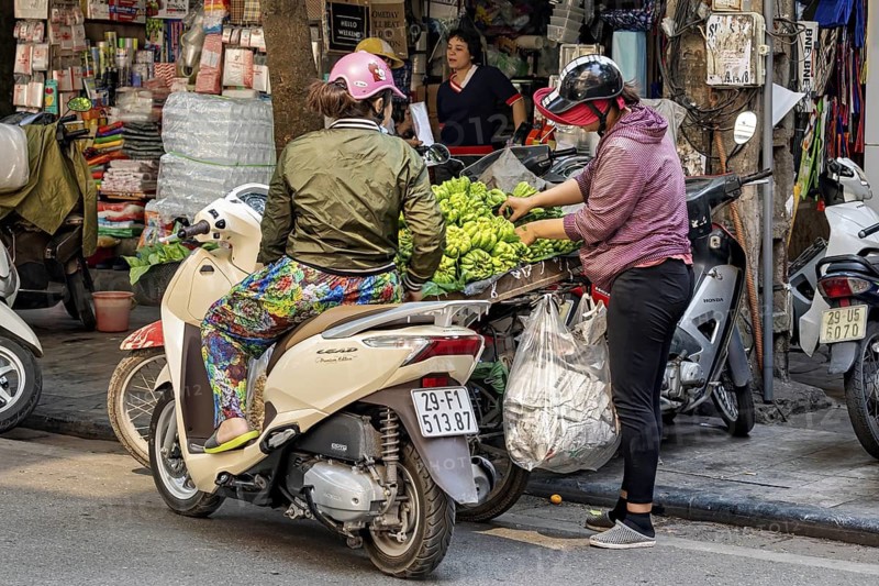 Two woman on scooters buy green fruit at a street stall in a busy city, woman selling fruit on the streets of Hanoi Vietnam