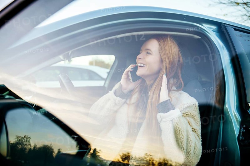 A beautiful pregnant girl with striking red hair is seated in her car, joyfully talking on her phone. The car window reflects the serene outdoor scenery