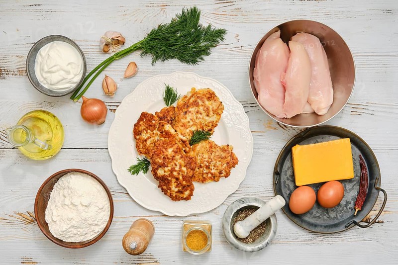 Raw ingredients surrounding plate with fried chicken patties. Raw chicken fillets, onion, eggs, flour, sour cream and spices on a tabletop. There is cooking oil and herbs nearby, ready for use