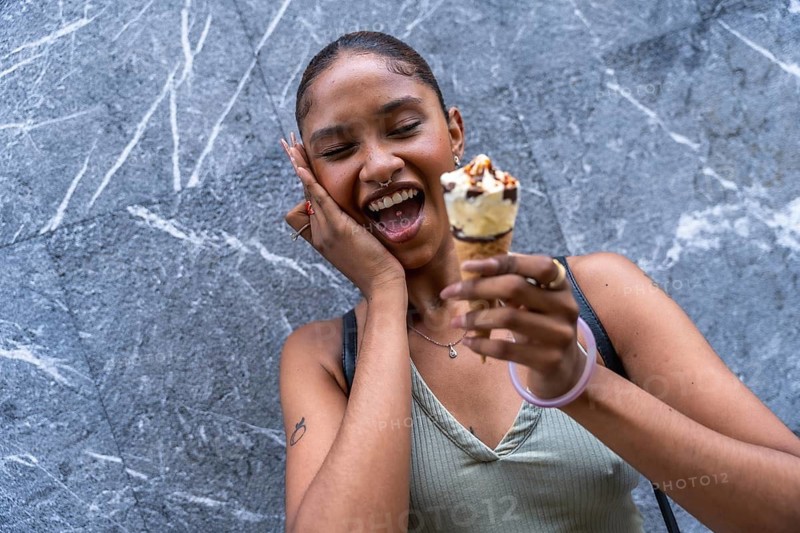 Young woman with dark skin and a nose piercing expressing joy and laughter, holding an ice cream cone in her hand against a grey marbled wall on a warm summer day, experiencing refreshing delight