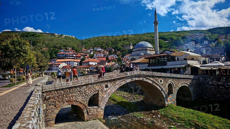 Urban scene with tourists on bridge over Prizren Bristrica river, mosque in background and hills under blue sky, Kosovo