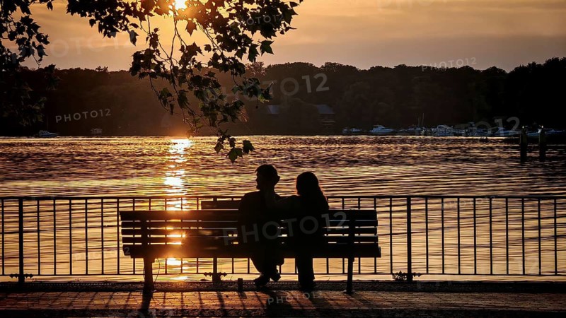 Silhouette in evening light, a couple sitting romantically on a bench and enjoying the sunset in warm colors by the water, Greenwichpromenade, Tegeler See, Tegel, Berlin