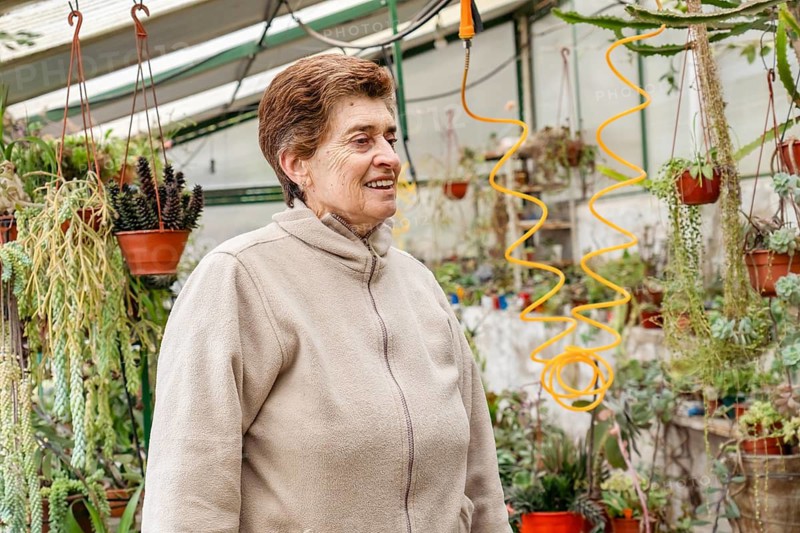Close-up portrait of a cheerful mature woman with short brown hair, smiling naturally inside her home greenhouse surrounded by hanging succulents and cacti