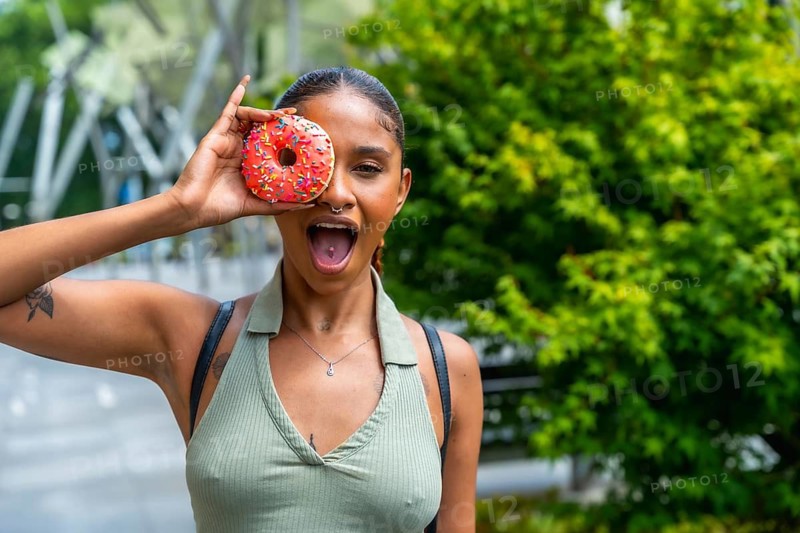 Young woman is having fun covering one eye with a colorful sprinkle donut and making a surprised face with her mouth open, enjoying a sweet moment outdoors