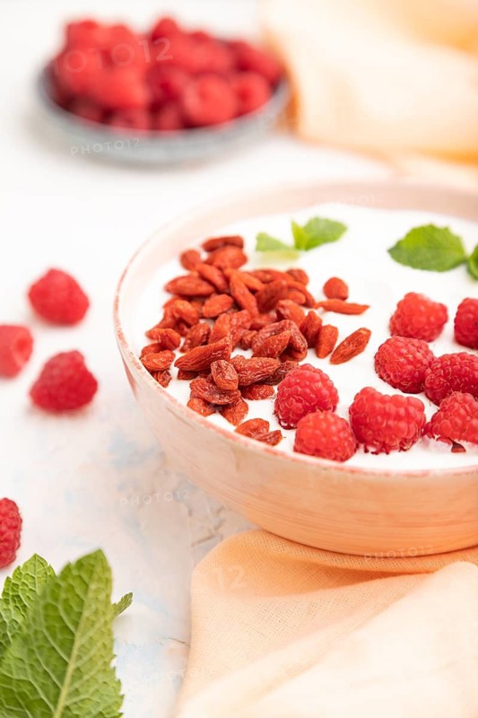 Yogurt with raspberry and goji berries in ceramic bowl on white concrete background and orange linen textile. Side view, close up, selective focus