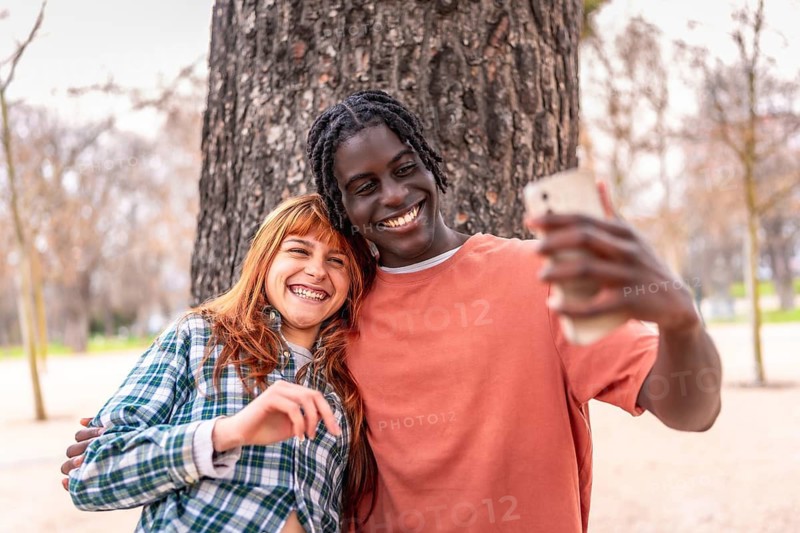 Young multiracial couple taking a selfie with a smartphone in a park, smiling and having fun