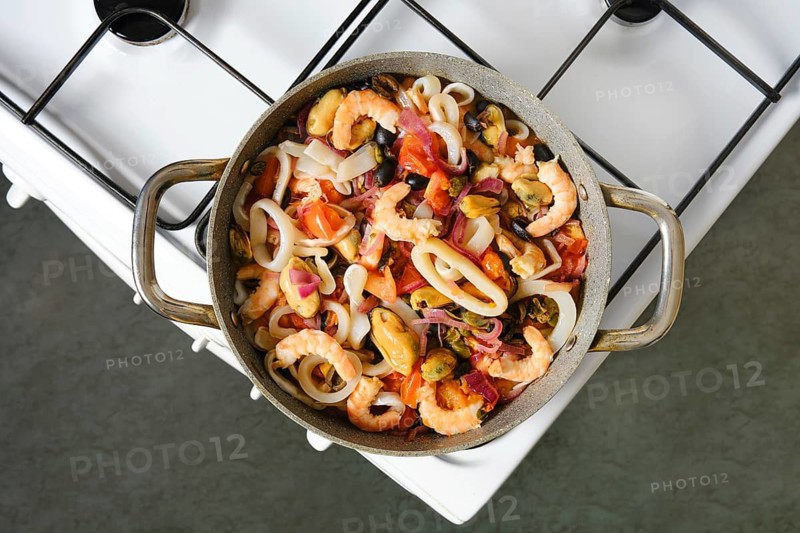 A pot holds a mixture of shrimp, mussels, and squid with chopped tomatoes and onions. Cooking seafood on a stove