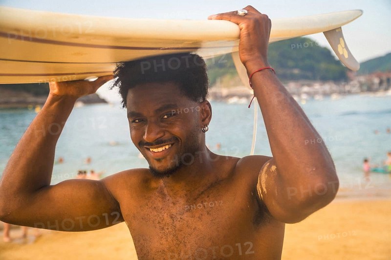 Young black man smiling at camera, carrying a surfboard on his head, standing on a sunny beach with the ocean and other people in the background, captured with a retro aesthetic