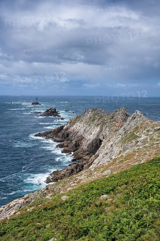Steeply sloping, rocky cliffs of the Breton coast under blue skies with light clouds. The view extends far across the rugged landscape of rocks and barren vegetation to the deep blue Atlantic Ocean, with the Phare de la Vieille lighthouse on a rocky island in the background. Waves break foaming on the rugged coastline under bright daylight. Pointe du Raz, Finistère, Brittany, France