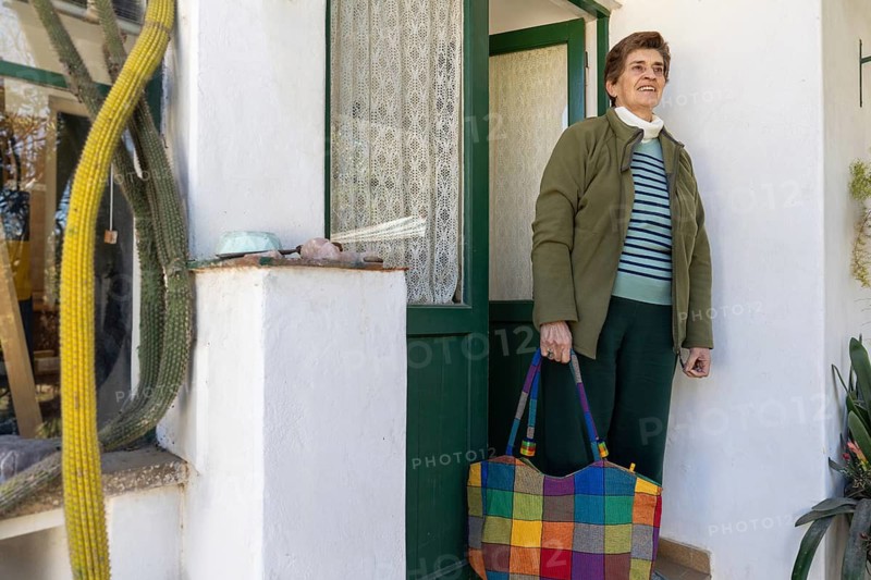 Older woman standing beside the entrance of her home with a colorful shopping bag, representing active aging and independent daily living