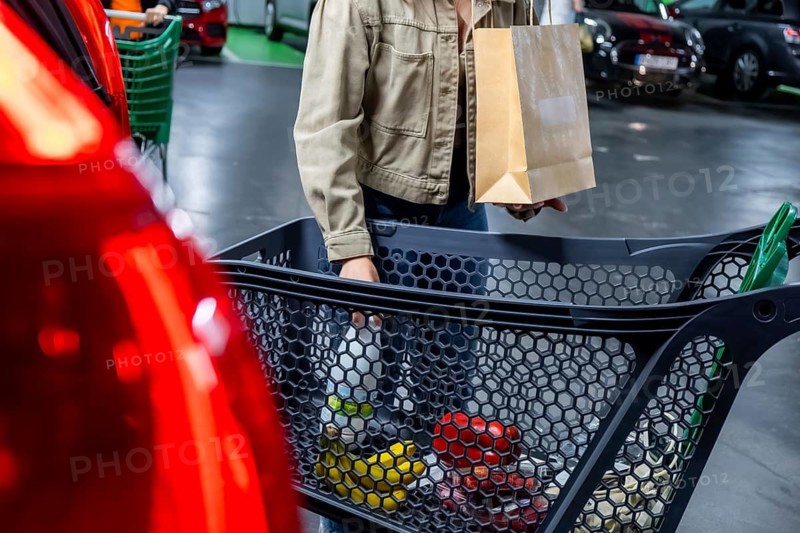 Customer shopping at a supermarket, carrying a paper shopping bag and pushing a cart full of groceries in a parking lot, representing daily errands and consumerism