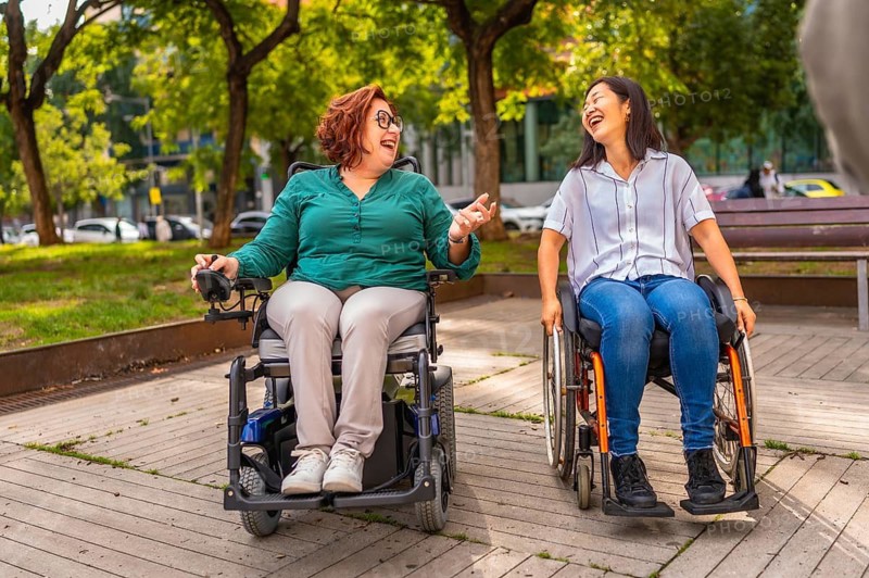 Two multiracial women with disability in wheelchair laughing talking together in a city park
