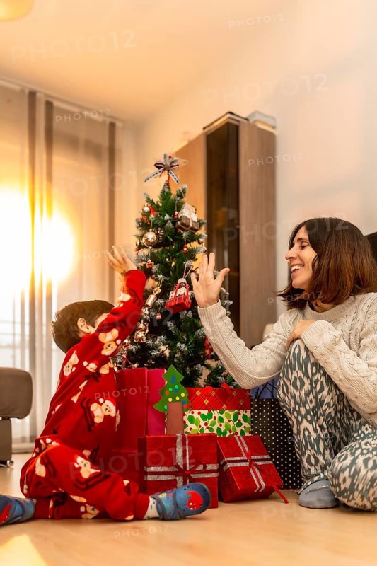 Happy mother and son bonding, wearing pajamas, and giving a high five next to a decorated christmas tree with wrapped gifts underneath, celebrating the holiday season