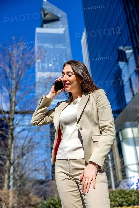 Businesswoman engaging in a phone call against a backdrop of modern skyscrapers and a clear blue sky, representing urban communication and professional connectivity