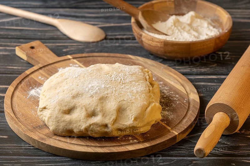 Fresh dough on a wooden board next to a bowl of flour and kitchen utensils