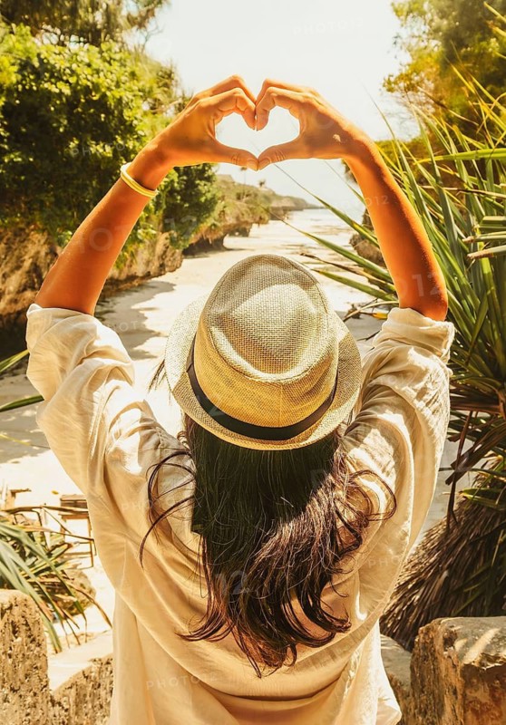 Young Woman tourist in straw hat framing heart sign with hands overhead looking down at tropical sandy cove beach surrounded by dramatic rocky cliffs vacation holiday lifestyle