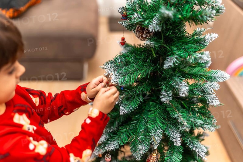 Young boy in red pajamas carefully hangs ornaments on a festive christmas tree at home, smiling with anticipation as family holiday decorating creates cozy, joyful memories
