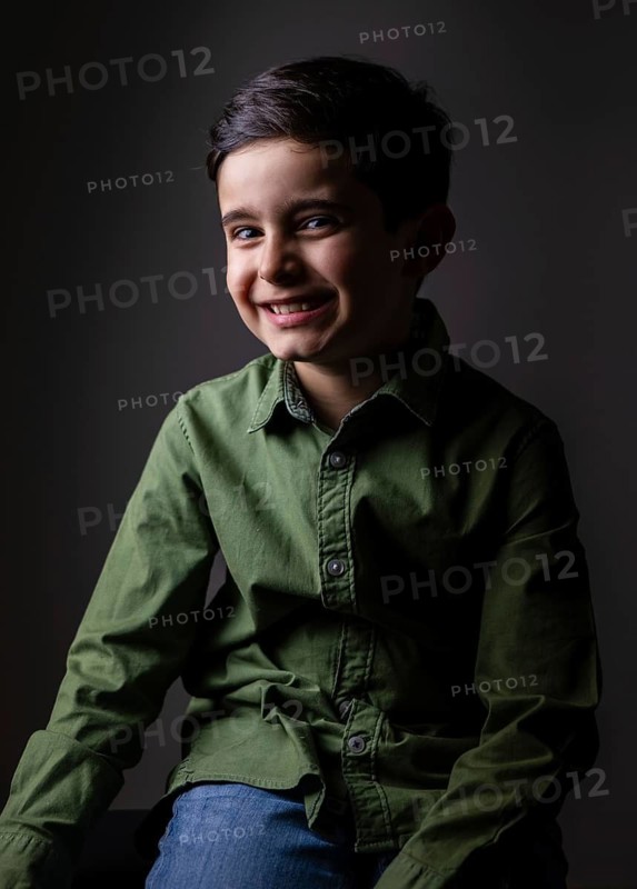 A young boy in a green shirt and jeans sits against a dark background, smiling warmly. The portrait captures his joyful expression and relaxed posture in soft lighting