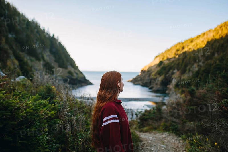 Side view of unrecognizable woman gazing at the serene waters of Shoe Cove Beach, surrounded by lush greenery, in Newfoundland, Canada. The calming ocean and rugged coastline create a picturesque scene