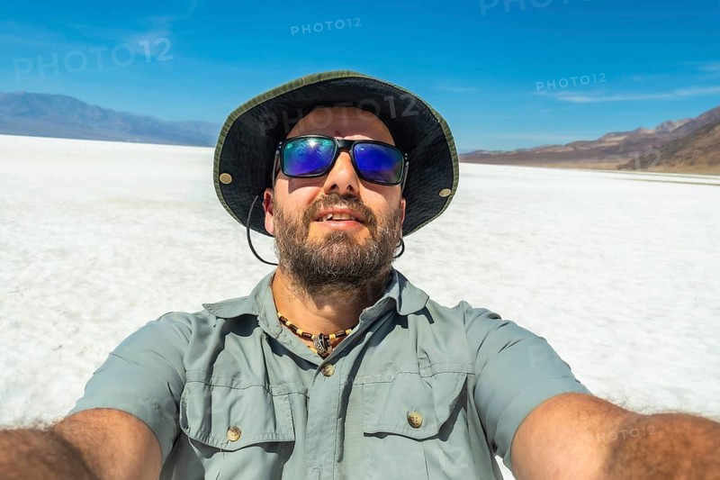 Man wearing a hat and sunglasses taking a selfie at badwater basin in death valley national park, exploring the vast, hot, and arid salt flats under a clear blue sky