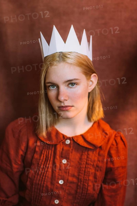 A young teenage girl with blonde hair wears a handmade paper crown and a vintage-style red dress. Her thoughtful gaze and warm lighting create an artistic and nostalgic portrait
