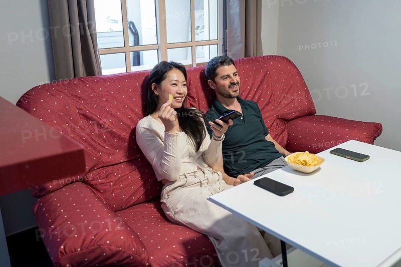 A diverse couple sits comfortably on a red couch at home, enjoying a relaxing day. They are sharing laughs and snacks, surrounded by a cozy and inviting atmosphere