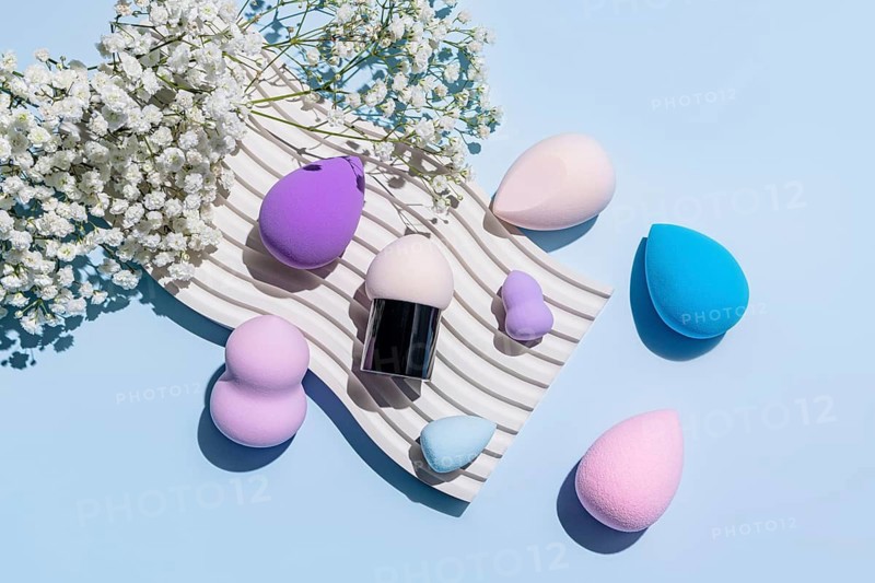 From above shot of various colorful makeup sponges, including beauty blenders, displayed elegantly on a textured mat alongside white blossoms on a pale blue background