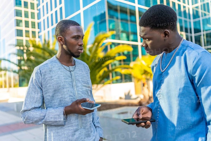Two senegalese businessmen wearing traditional dashiki attire are using their smartphones in front of a modern office building