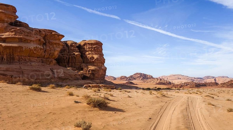 The Wadi Rum desert in Jordan, showcasing sandy terrain, rocky formations, and a clear blue sky. A dirt path leads through the mesmerizing landscape, surrounded by majestic red rock mountains that echo the rich history of this ancient area