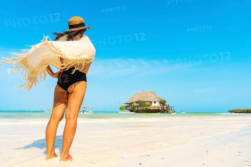 Woman tourist wear bohemian tunic walks across white sandbar toward the iconic Rock Restaurant in Zanzibar. Low-tide coastal scene with turquoise water and blue sky