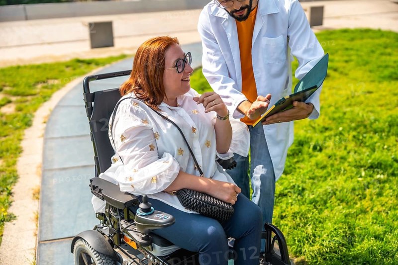 Doctor showing medical information to a smiling woman sitting in a wheelchair in a park, promoting accessibility and healthcare support