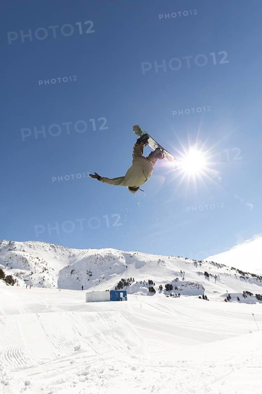 A snowboarder performs a daring aerial trick against a stunning mountain backdrop under a clear blue sky. The untouched snow suggests a perfect day for backcountry adventure