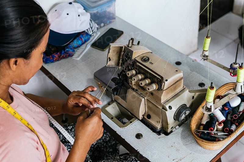 A asian seamstress operates a sewing machine in a textile workshop, focusing on detail. Nearby are spools of thread in a basket. The setting is an artisan's workspace